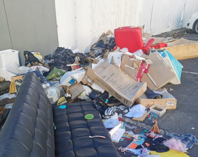 A dumped black couch, cardboard boxes and bagged household rubbish piled beside a wall
