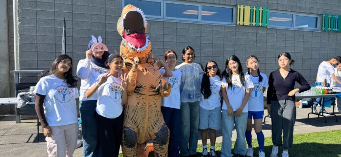 Youth Project NZ volunteers in branded t-shirts at an outdoor community event, with a person in an inflatable dinosaur costume