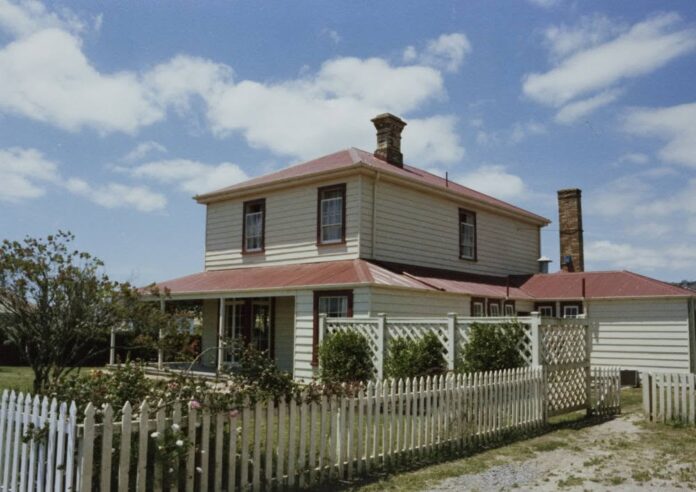 Two-storey white weatherboard house with a red roof, verandah and brick chimneys, behind a white picket fence