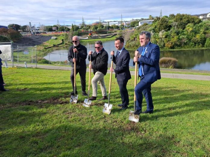Four men in suits hold ceremonial spades on grass beside a lake at the Eastern Busway sod-turning, with construction fencing and the Botany skyline behind