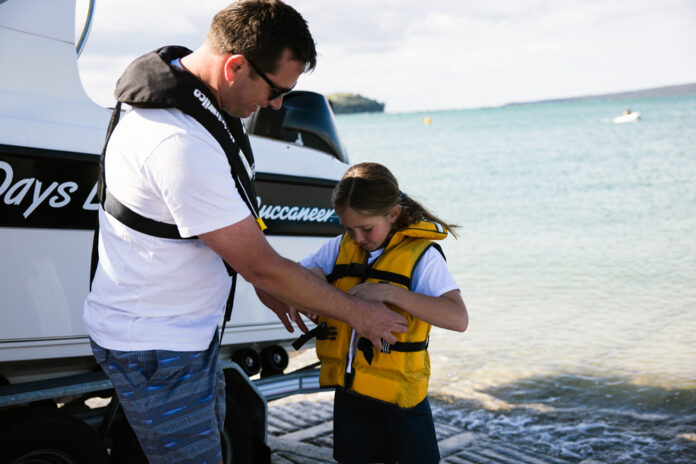 A man fits a yellow lifejacket onto a child beside a boat at the water's edge