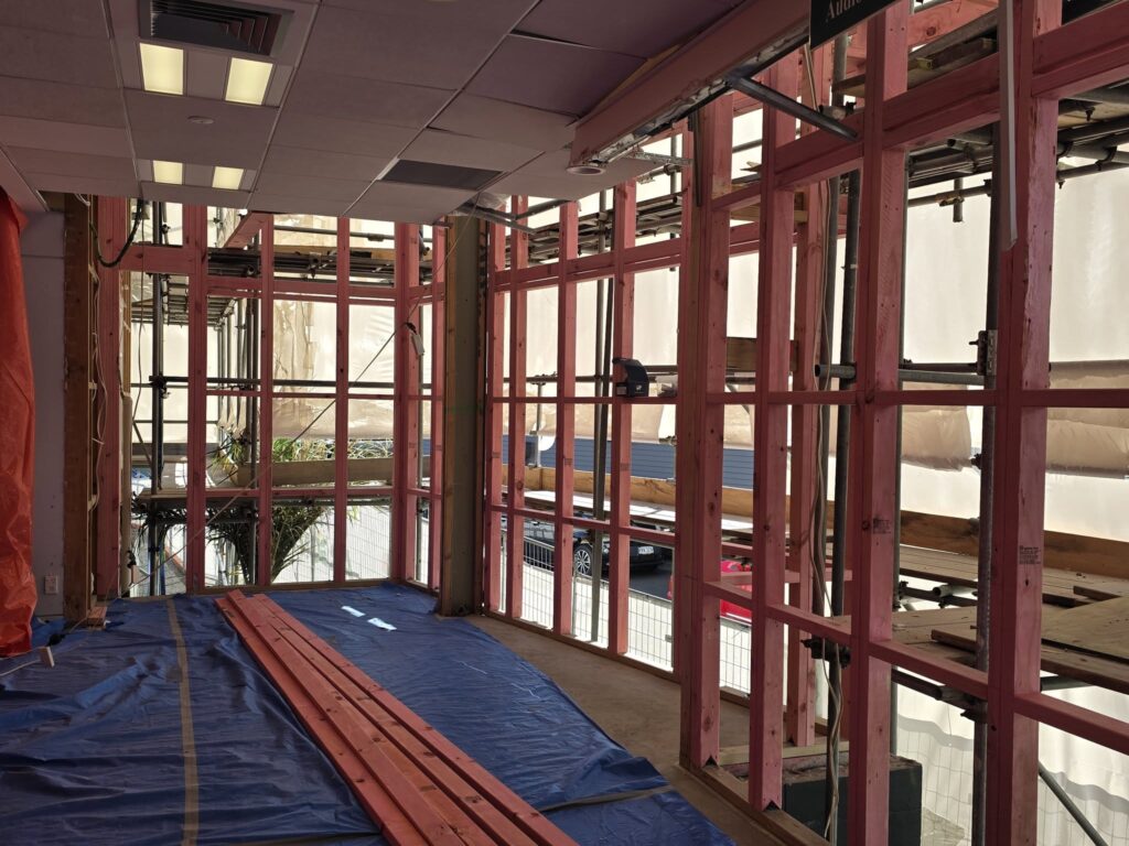 Interior of Howick Library during renovation, showing exposed timber wall framing where cladding has been removed, a tarpaulin-covered floor and stacked timber