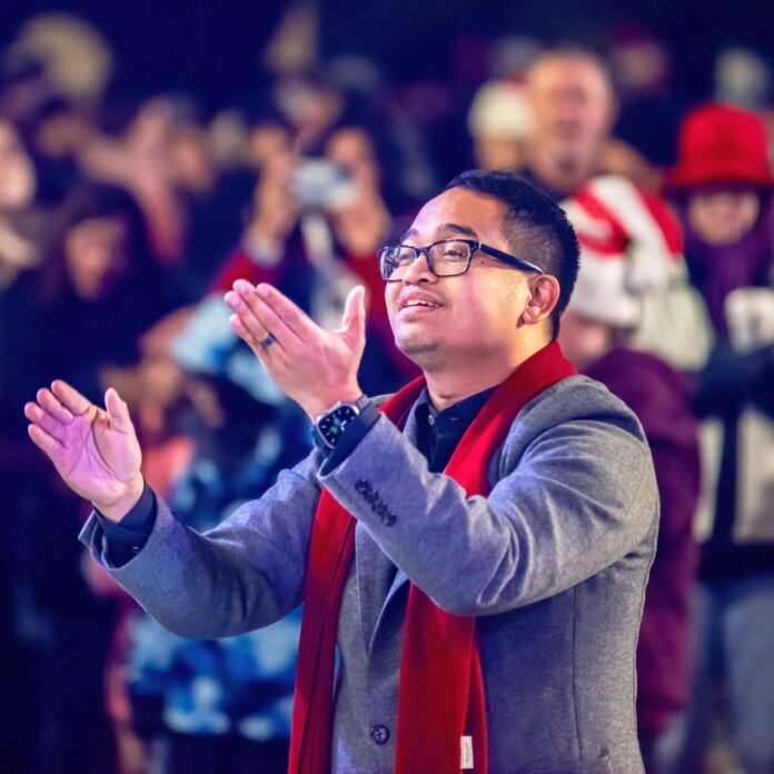 Conductor Jason Sabino, in glasses and a red scarf, conducts with both arms raised in front of a crowd
