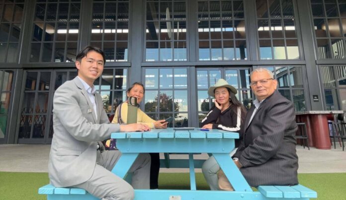 Four people seated at a turquoise picnic table outside the glazed frontage of Flat Bush Library in Ormiston, with Kai Zeng at the left of the group and Yuva Adhikary at the right