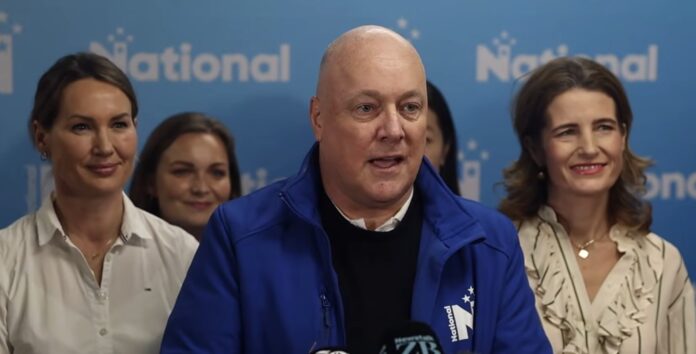 Christopher Luxon speaks at a lectern in front of a blue National Party backdrop, with four women standing behind and beside him.