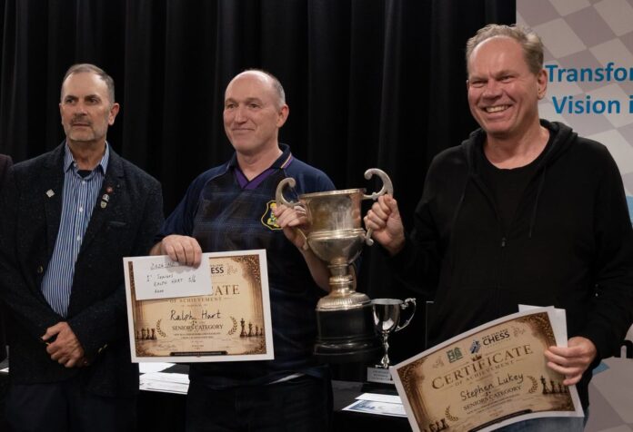 Three men at a chess prizegiving in front of a black curtain, two holding certificates and one holding a large silver trophy.