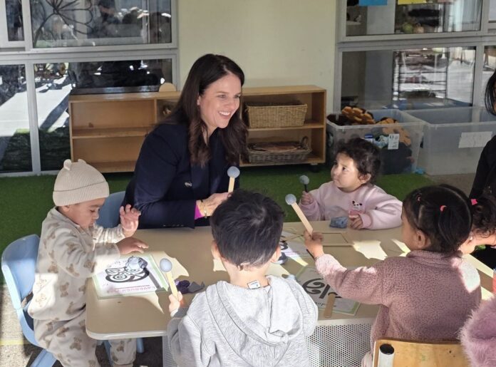 Education Minister Erica Stanford sits at a low table with toddlers at an early childhood centre, holding wooden rhythm sticks during a group activity.