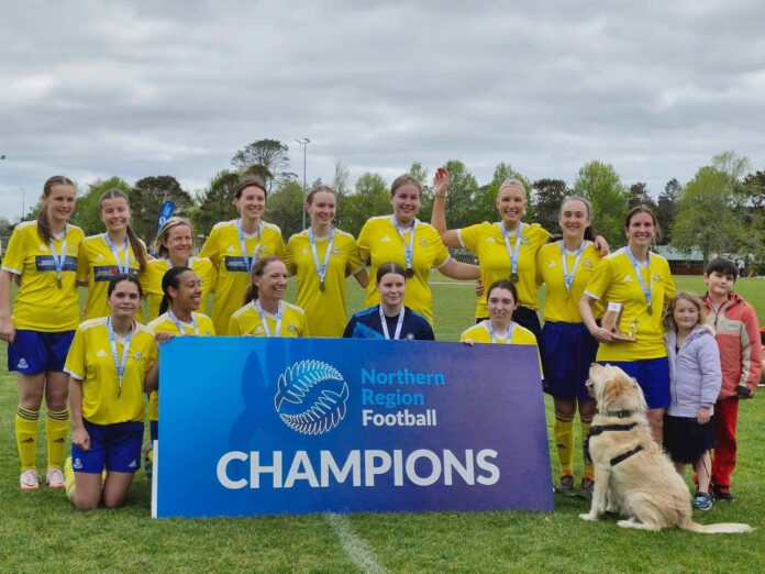 A women's football team in yellow and blue strip pose with medals and a trophy behind a Northern Region Football champions board on a grass field.