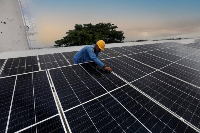 A worker in a hard hat crouches to adjust a rooftop array of solar panels at dusk.