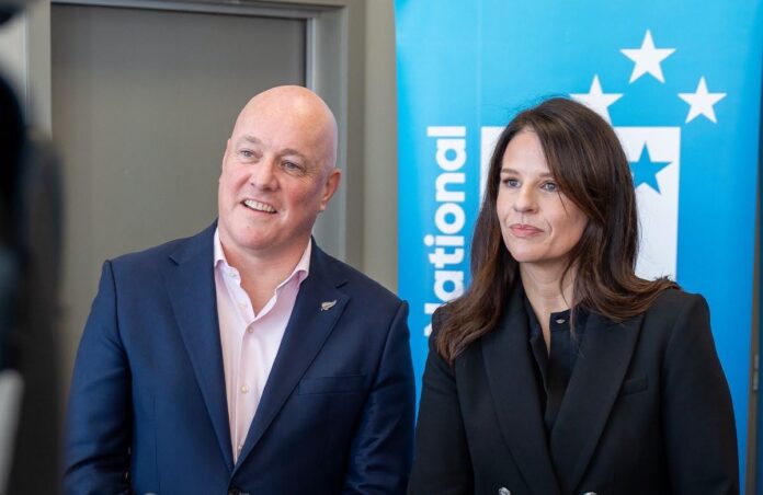 Christopher Luxon and Erica Stanford stand side by side in front of a blue National Party banner.