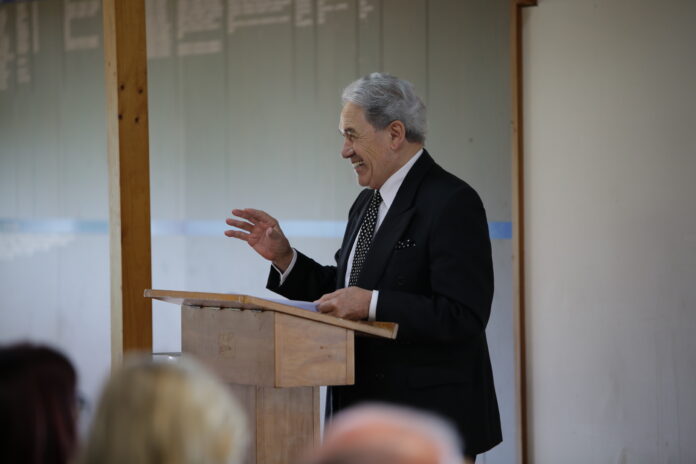 Winston Peters speaking from a wooden lectern at a Grey Power meeting, right hand raised mid-gesture, audience heads in the foreground.