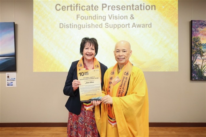 Adele White in a yellow Art Salon sash holds a framed certificate beside a Buddhist nun in saffron robes, in front of a screen reading Certificate Presentation.