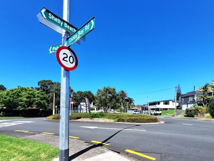Street name signs for Shelly Beach, Cockle Bay Road and Pah Road on a pole above a 20km/h speed limit sign, at a suburban intersection under a clear sky.