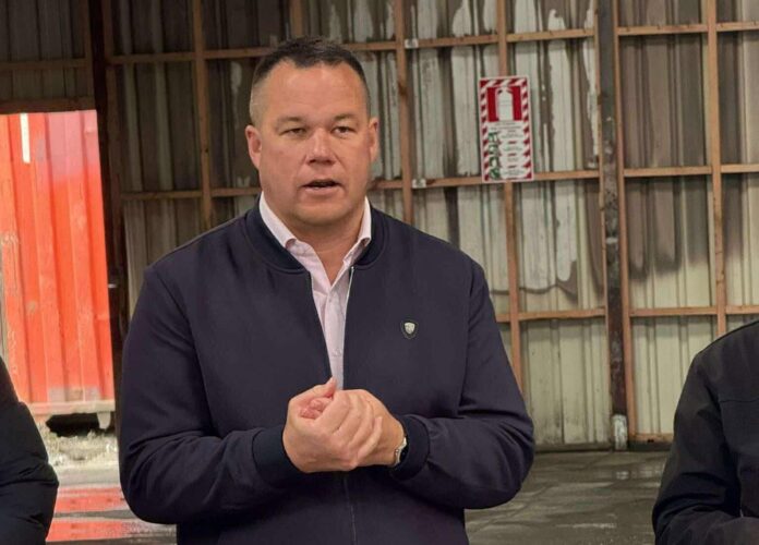 Daniel Newman speaking with his hands clasped, in an industrial shed with corrugated walls and a shipping container behind him.