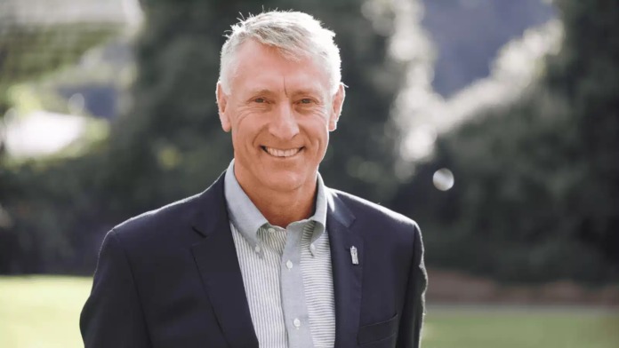 Craig Stobo in a dark suit jacket and pale striped open-necked shirt, smiling at the camera outdoors with blurred trees behind him.