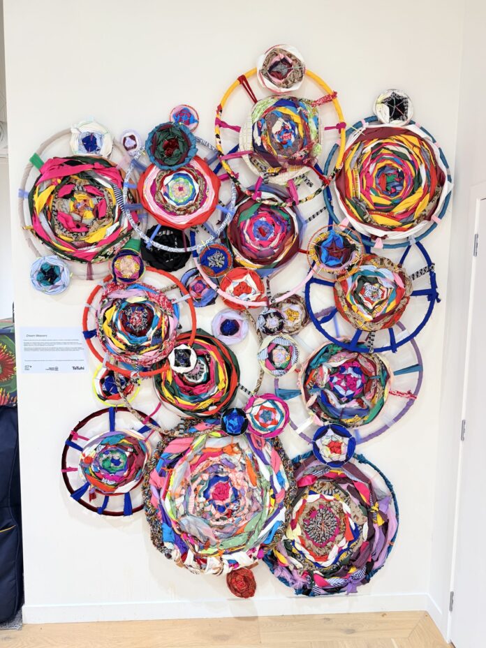 A large wall installation of about 30 circular hoops of different sizes, each woven with brightly coloured fabric strips, mounted on a white wall above a wooden floor.