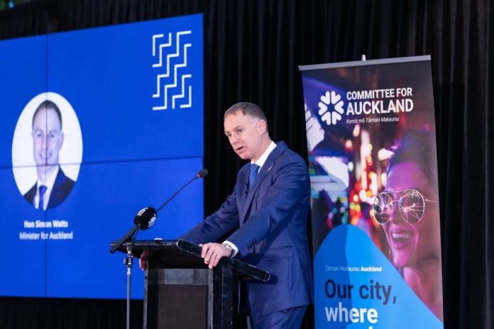 Local Government Minister Simon Watts speaking at a lectern, with his name displayed on a screen behind him.