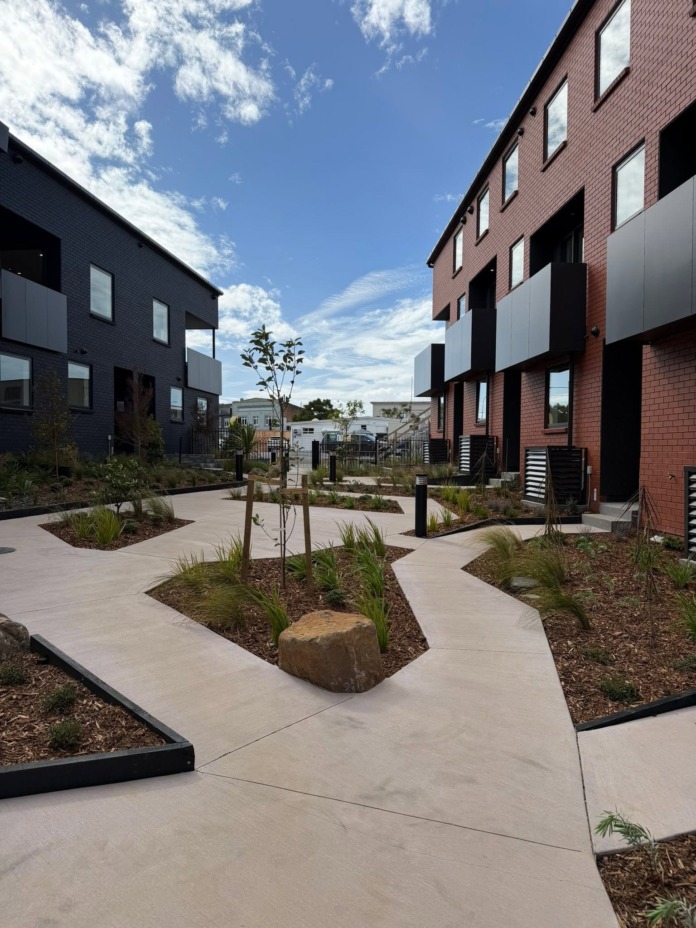 A landscaped courtyard between two three-storey terraced apartment blocks, one charcoal brick and one red brick.
