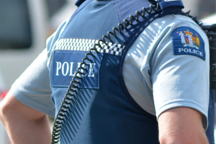 Close-up of a New Zealand Police officer's shoulder and stab vest showing the POLICE badge and chequered band, with the officer's face out of frame.