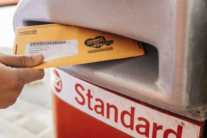 A hand posts an Auckland Council voting envelope into an NZ Post box