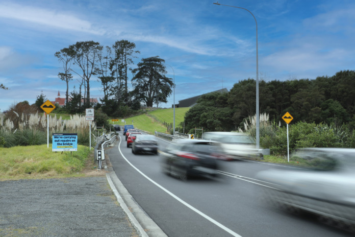 Cars moving along Chapel Road, Flat Bush, past grass verges with no footpath. A roadside sign advertises Auckland Transport bridge repairs. Two churches stand on the skyline.
