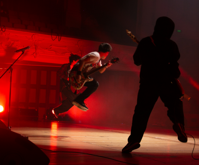 A teenage guitarist leaps into the air on a red-lit stage as a bassist in silhouette plays in the foreground.