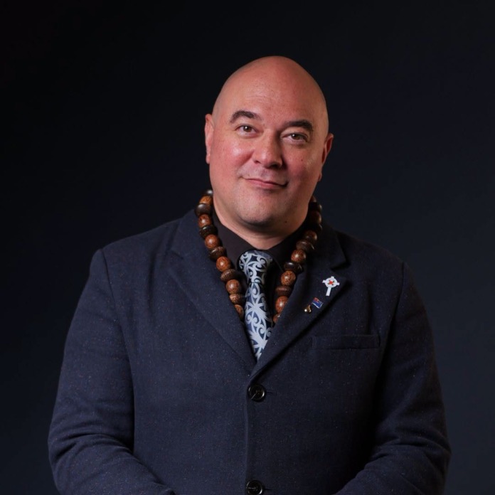 Elliot Ikilei, New Zealand First's candidate for Takanini, in a dark jacket and wooden bead necklace against a black studio background.
