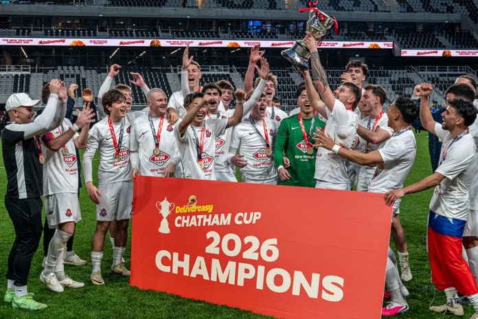 Fencibles United players and staff celebrate on the pitch, one player holding the Chatham Cup above his head, behind a board reading Chatham Cup 2026 Champions.