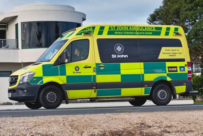 A yellow and green Hato Hone St John ambulance on a seafront road, with a white curved-fronted house and a pohutukawa tree behind it.