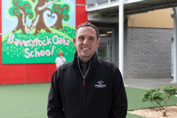 Ian Dickinson standing outside Baverstock Oaks School, in front of a painted sign carrying the school's name.