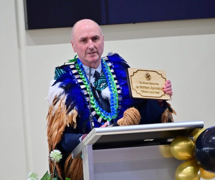 Ian Morrison, wearing a feathered korowai, speaks at a lectern holding a wooden plaque reading Ian Morrison Gymnasium.