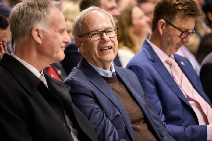 An older man in a navy jacket and brown jumper smiles while seated in an audience between two other men.