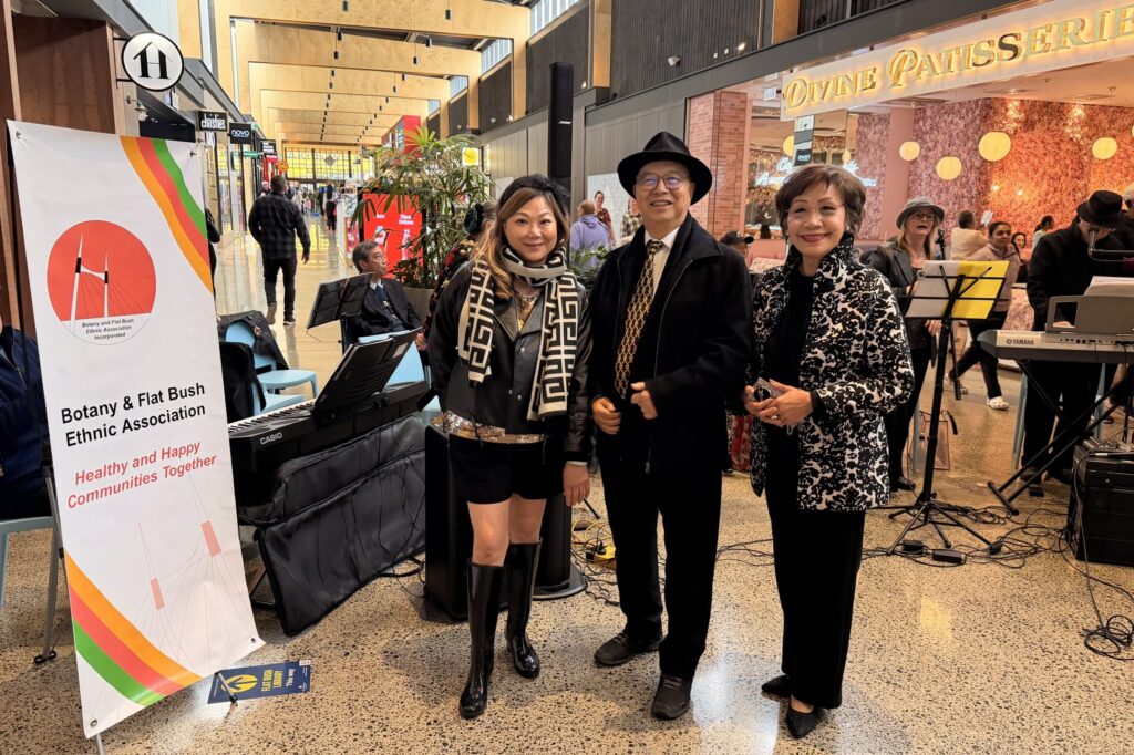 Peter Young in a black hat and coat stands between two women, one of them association deputy chair Eleanor Mak, beside the association's Healthy and Happy Communities Together banner, with keyboards and music stands behind them in the Ormiston Town Centre mall.