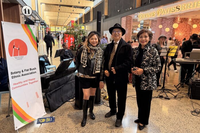Peter Young in a black hat and coat stands between two women, one of them association deputy chair Eleanor Mak, beside the association's Healthy and Happy Communities Together banner, with keyboards and music stands behind them in the Ormiston Town Centre mall.