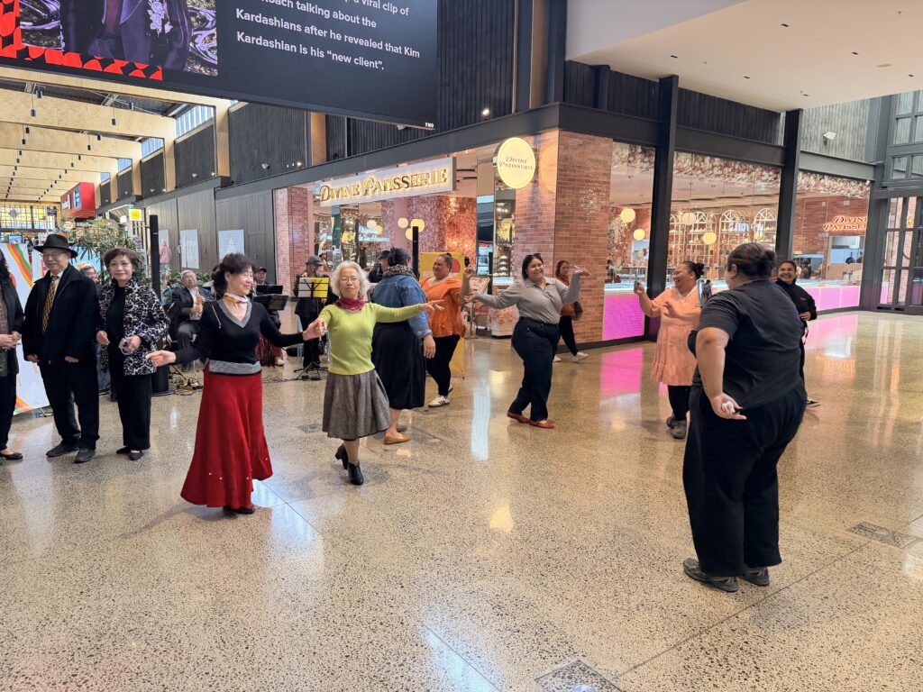 Dancers, most of them older women, hold hands in a line across the polished floor of the Ormiston Town Centre mall, with a man in a black hat and a woman in a patterned jacket watching from the left and shoppers behind.