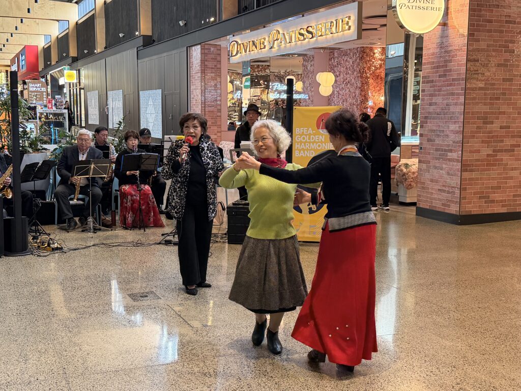 Two older women, one in a green jumper and grey skirt and one in a long red skirt, dance together in the mall while a woman in a patterned jacket sings into a red microphone, with seated band members and a yellow Golden Harmonies Band banner behind them.