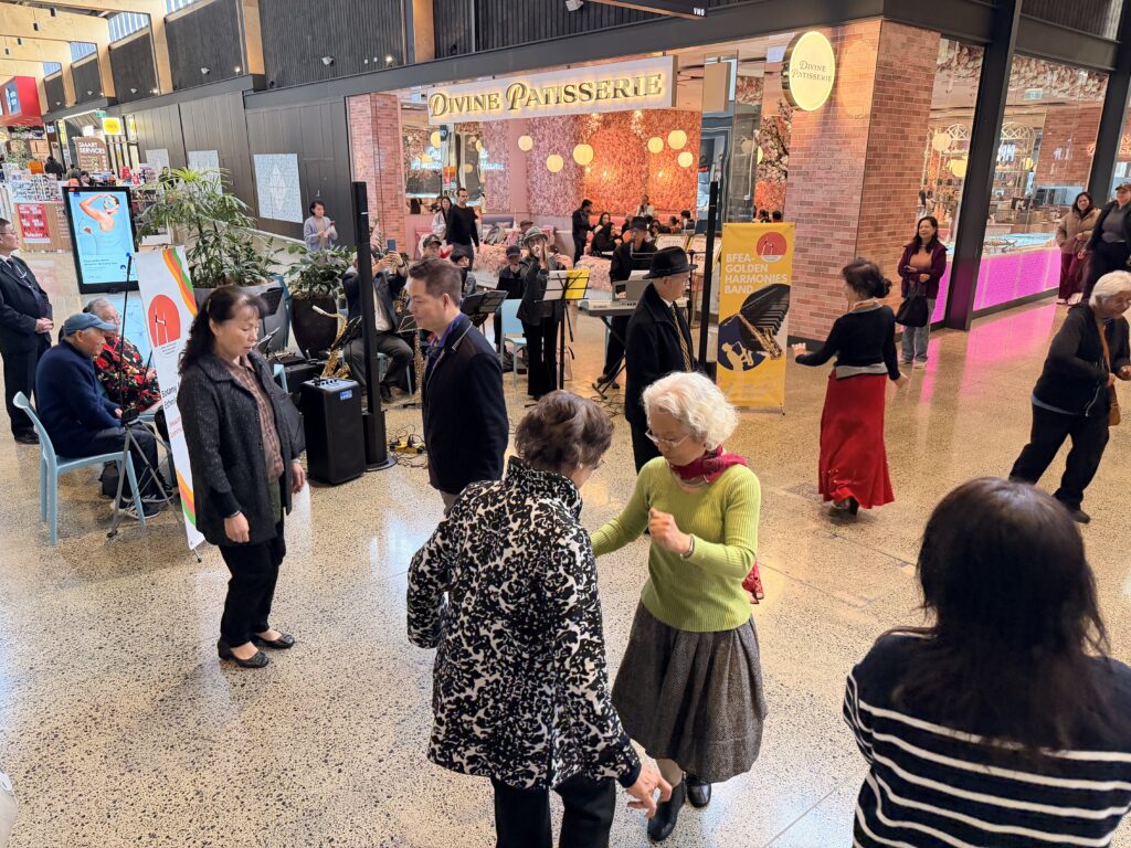 An older woman in a green jumper dances with a woman in a black-and-white patterned jacket in the mall, as others dance and watch around them in front of seated band members, a man in a black hat and a yellow Golden Harmonies Band banner.