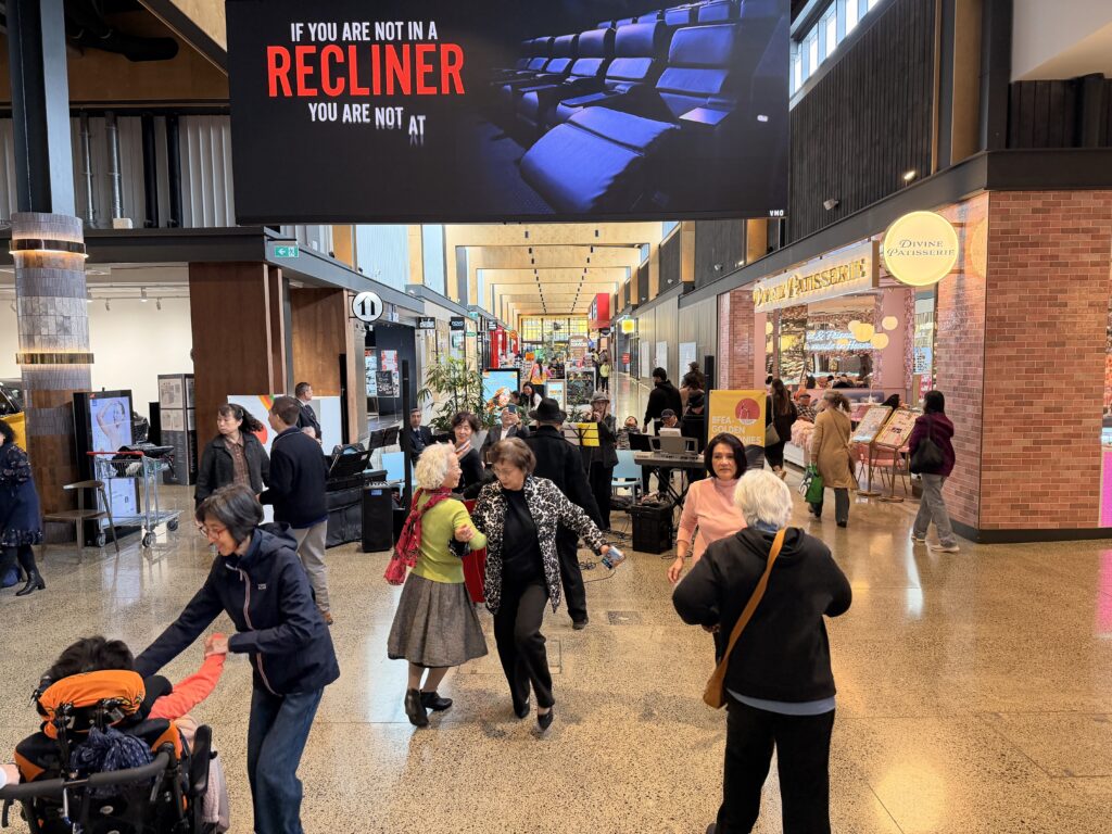 Dancers and shoppers mingle in the wide mall at Ormiston Town Centre, where a woman takes the hand of a person in a wheelchair to dance, and the Golden Harmonies Band plays in the background.