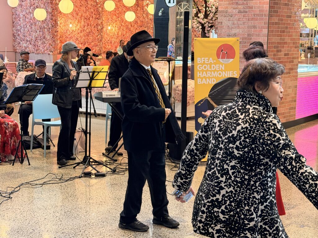 Peter Young, in a black hat, long black coat and patterned gold tie, smiles in front of the Golden Harmonies Band and its yellow banner, as a woman in a black-and-white patterned jacket dances past in the foreground.