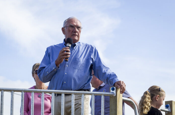 Malcolm Bell, in a blue shirt, speaks into a microphone from a raised wooden platform with a metal railing, under a blue sky.