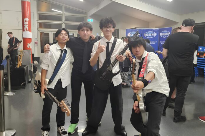 Four teenage musicians in white shirts and black ties pose backstage with a bass and two electric guitars, in front of Rockshop banners.