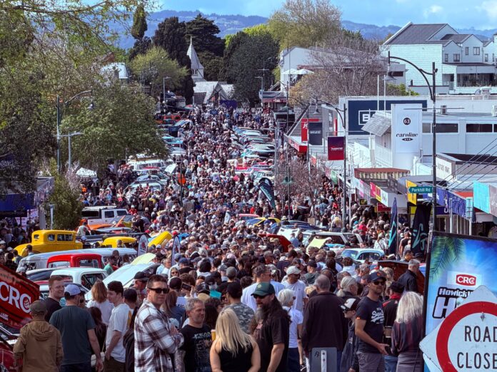 A dense crowd fills a Howick village street lined with classic cars and shopfronts, behind a red road-closed sign, cones and a CRC Howick Village HOP banner.