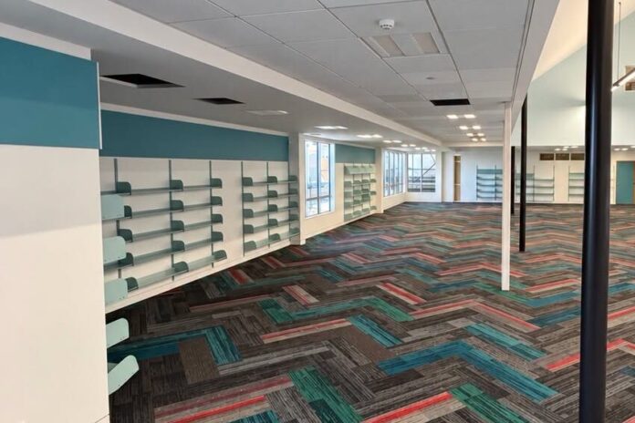 Inside the refurbished Howick Library, a long open room with teal feature walls, empty wall shelving, and grey, teal and red herringbone carpet