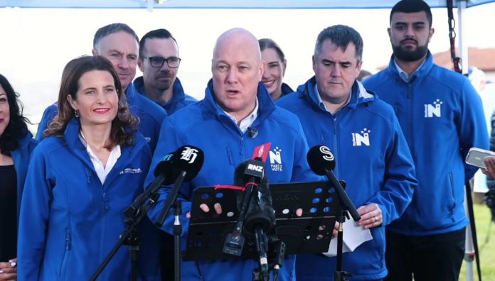 Christopher Luxon speaks at a lectern crowded with media microphones, with Nicola Willis to his left, Chris Bishop to his right and National supporters in blue party jackets behind them.