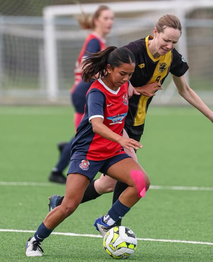 Mikaela Bangalan, in a red and navy kit, drives forward with the ball as an opponent in black and yellow chases.