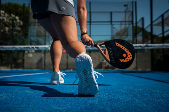 A padel player seen from behind on a blue court, holding a black and orange racket, with the net and wire fencing beyond.