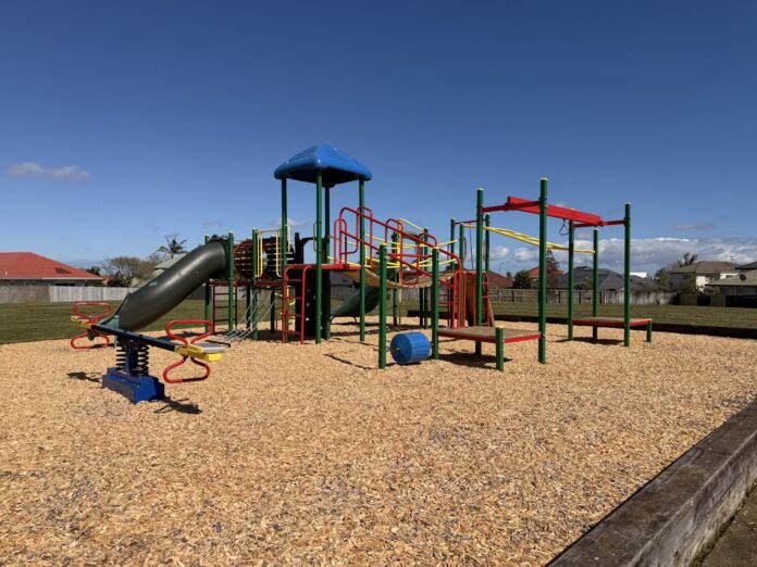A green, red and blue playground with a tube slide, climbing frames, monkey bars and a seesaw on bark chip at Tarnica Park, with houses behind under a blue sky.