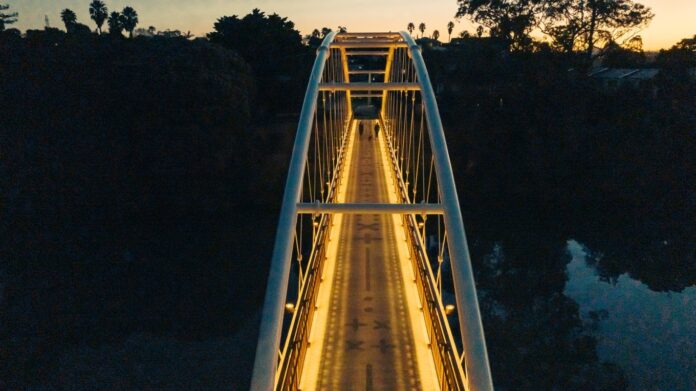 Aerial view at dusk of the Te Kopua o Hiku footbridge over the Panmure Basin, its arched white frame and patterned deck lit gold, with two people walking across.